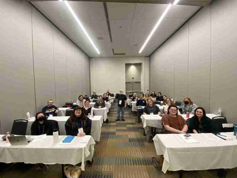 A picture of Jim Guy, standing in the middle of a conference room with dozens of attendees, all sitting at tables, smiling at the camera